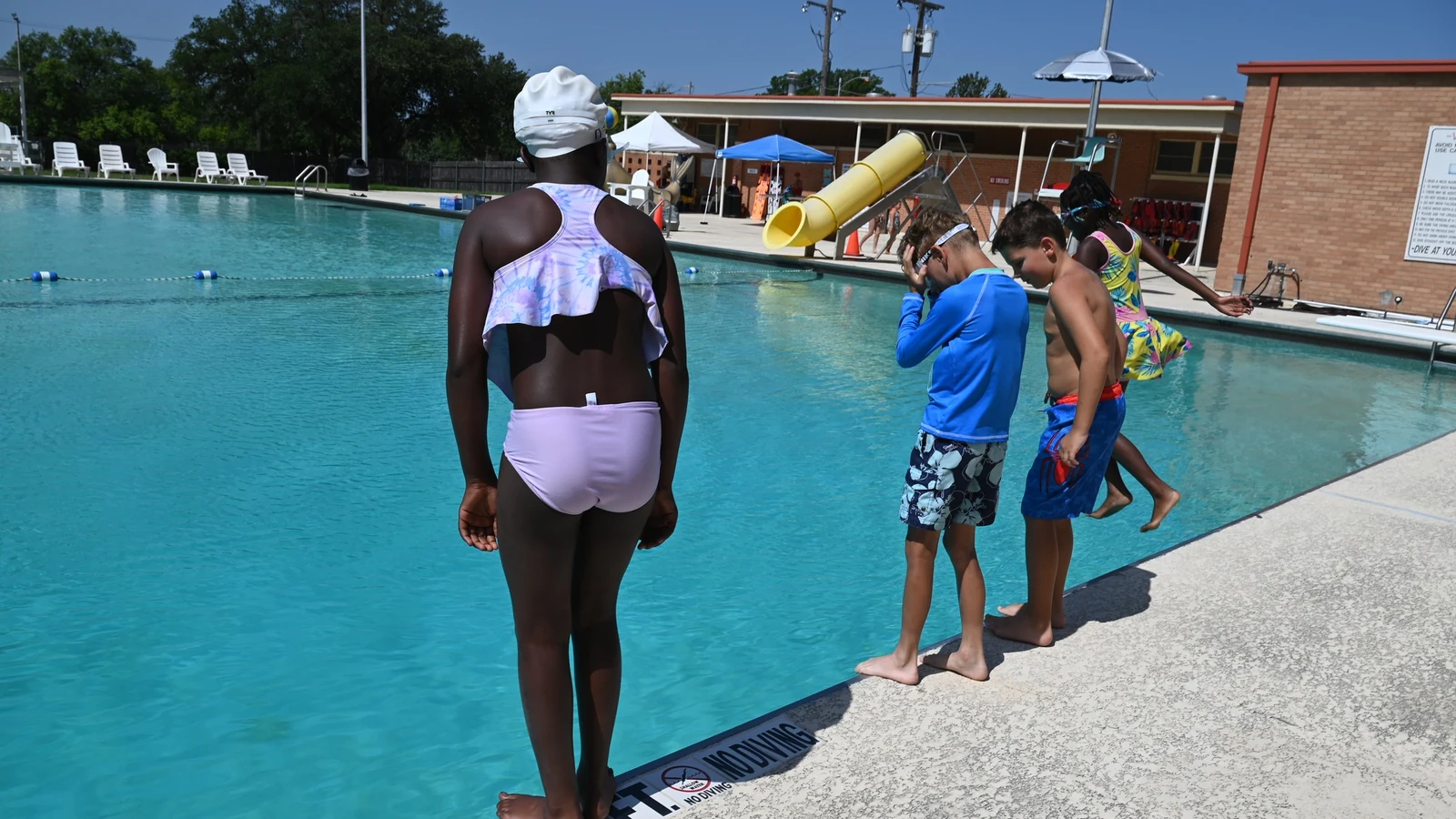 Children stand at the edge of an outdoor pool during a supervised swimming lesson.