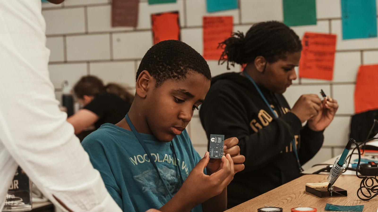 Students assemble small circuit boards at tables while an instructor helps them.