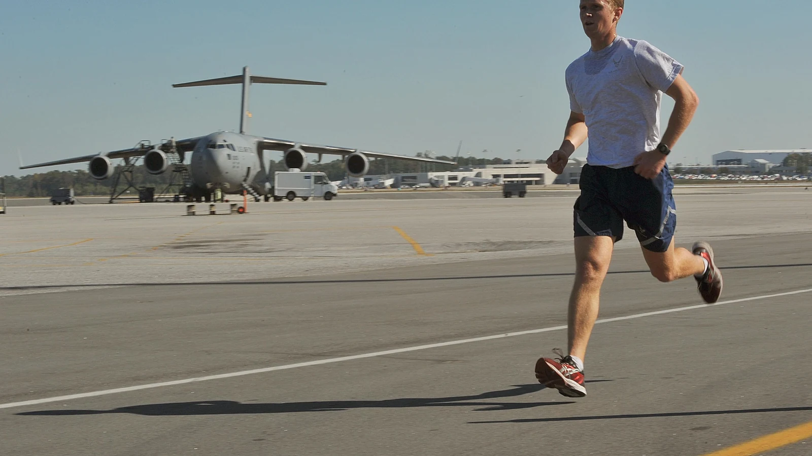 A runner in a red shirt strides down a broad runway with a large military transport aircraft behind him.