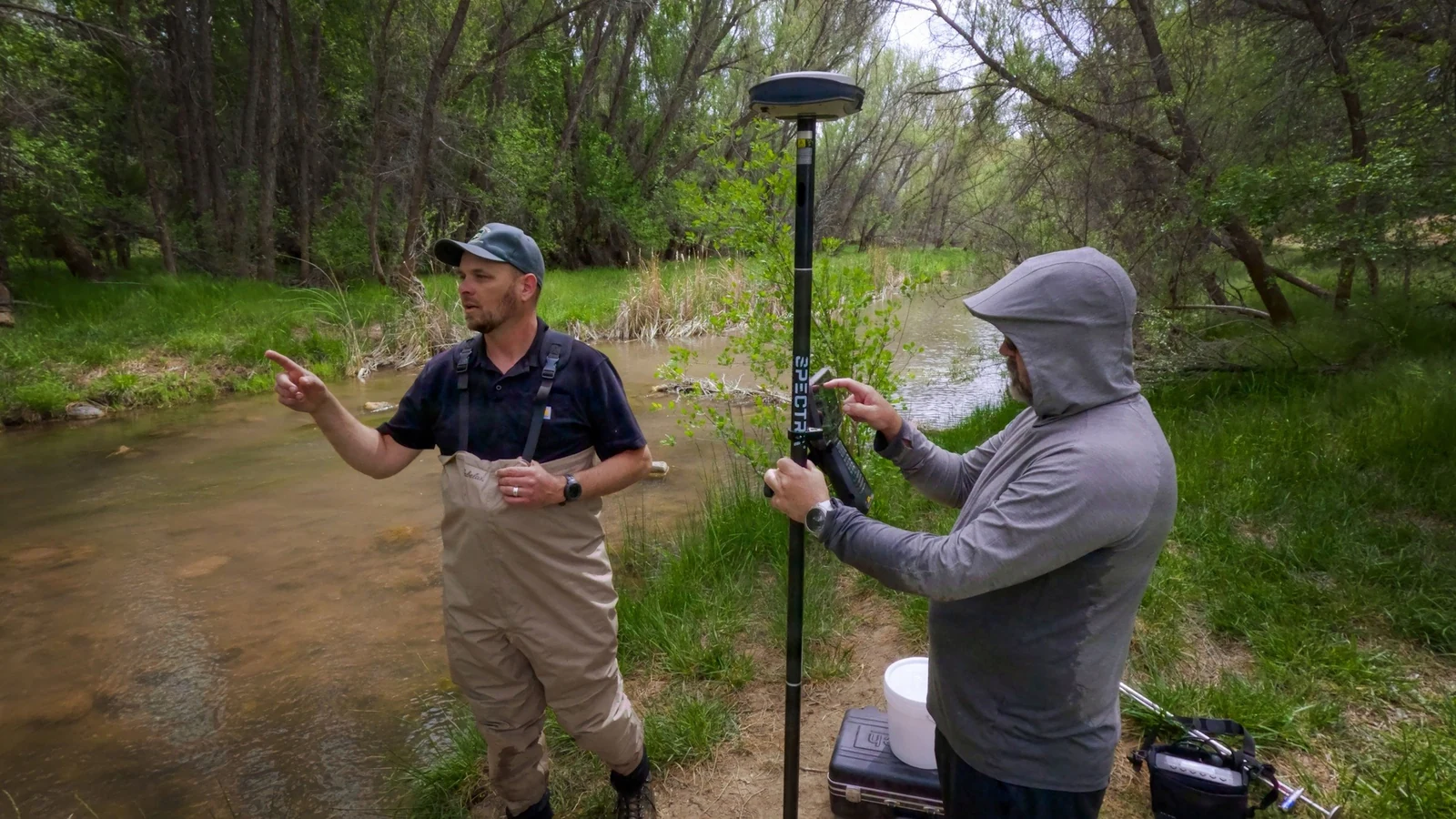 Two water researchers stand on a riverbank beside monitoring equipment.