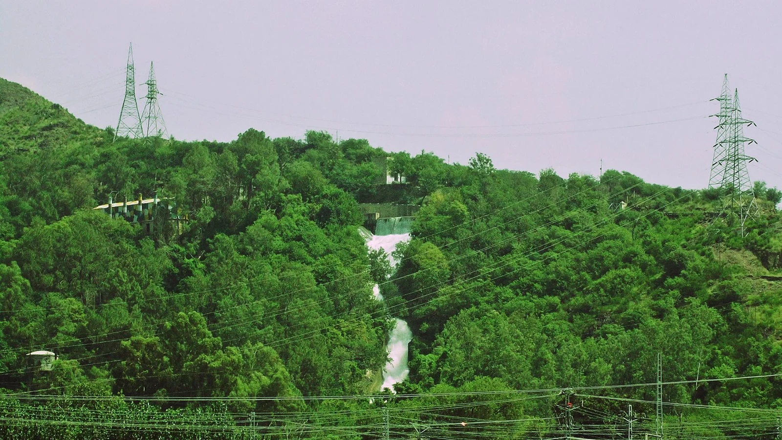 Steel electricity pylons and power lines cross a green hillside above a waterfall in Malakand, Pakistan.