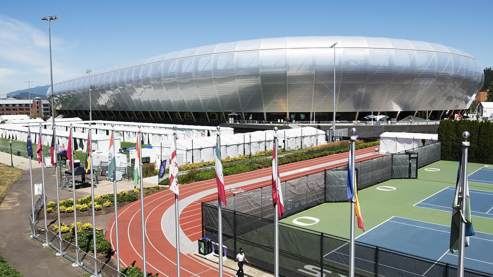 Hayward Field’s silver stadium shell rises above a red track, international flags and event tents under a clear blue sky.